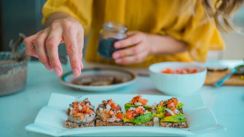 Hand sprinkling seeds on avocado toast with diced tomatoes, featuring a fresh and healthy dining setup.